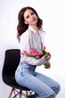 Studio portrait of a beautiful sexy slim young caucasian brunette in white shirt and blue jeans sitting with a bouquet of pink tulips