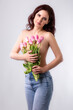 Studio portrait of a beautiful sexy slim young caucasian brunette standing topless and in blue jeans with a bouquet of pink tulips in her hands covering her breasts