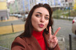 © Nick ALDI - Charming lady in a brown autumn coat takes a selfie on the street with a peace sign