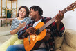 © Mediteraneo - Happy young couple singing a song playing guitar on sofa in living room at home