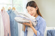 © KMPZZZ - Feel softness, hygiene. Smile asian young woman touch fluffy white shirt smelling fresh clean pile, stack clothes, comfort sniff after washing laundry. Household work at home, chore of maid concept.