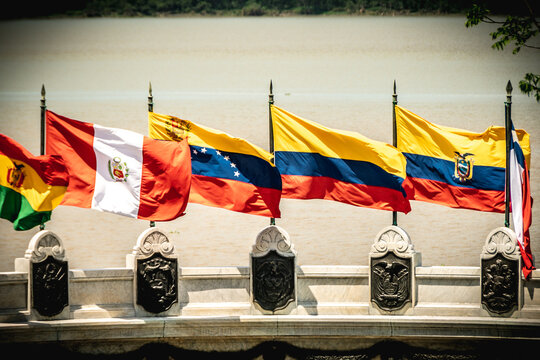 A close-up photo of a row of Latin American country flags flying in front of a body of water. The flags are from Peru, Bolivia, Venezuela, Ecuador and Colombia. They are moving with the wind. 