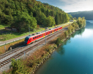 Wall Mural - Aerial view of red modern high speed train moving near river in alpine mountains at sunset in autumn. Top view of train, rural railroad, lake, road, green trees in fall. Railway station in Slovenia