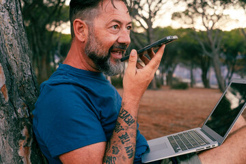 Wall Mural - One happy man do voice phone call outdoor at the park with computer on his legs sitting on the ground. Communication and outdoor connection. Travel digital nomad and open air office concept lifestyle