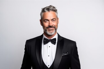 Wall Mural - Handsome mature man in tuxedo looking at camera and smiling while standing against white background