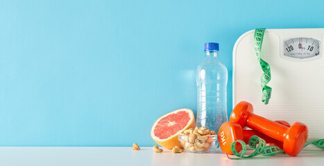 Wall Mural - Wholesome living depiction: side view of table decorated with scale, measuring tape, dumbbells, bottle of water, cashew nuts in bowl, and half of grapefruit, set against blue wall with space for text