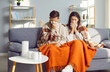 © Studio Romantic - Young couple covered in blankets sitting on sofa at home with seasonal flu or cold. Sick unhappy man with woman sneezing into a tissue suffering from runny nose and feeling unhealthy with influenza.