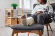 Cropped view of woman with leg in plaster bandage and crutch sittingin armchair at home. Selective focus of sitting African American woman with broken leg. Treatment at home, recovery concept