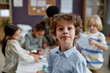 Portrait of cute curly haired boy looking at camera while enjoying arts and crafts class in preschool, copy space