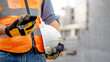 © Summer Paradive - Construction worker man with orange reflective vest holding white protective safety helmet using smartphone at unfinished building site. Male engineer or foreman stay connected with social media app