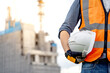 © Summer Paradive - Safety workwear concept. Male hand holding white safety helmet or hard hat. Construction worker man with reflective orange vest and protective gloves standing at unfinished building with tower crane