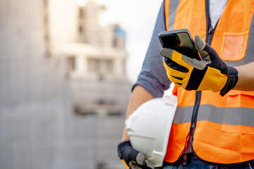 Construction worker man with orange reflective vest holding white protective safety helmet using smartphone at unfinished building site. Male engineer or foreman stay connected with social media app