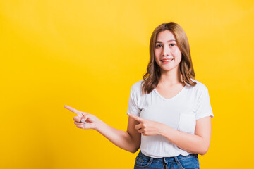 Wall Mural - Asian Thai happy portrait beautiful cute young woman standing wear white t-shirt pointing finger away side looking to camera, studio shot isolated on yellow background with copy space