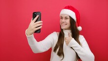 Cute Young Woman Wear White Knitted Sweater And Santa Hat Posing Doing Selfie Shot On Mobile Cell Phone Show Thumb Up, Isolated On Pastel Red Background In Studio. Happy New Year 2024 Holiday Concept