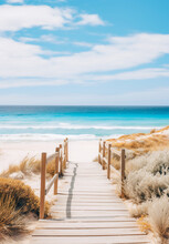 A Wooden Boardwalk With Wooden Railings Leading Through The Sand Dunes To A Sandy Beach With Clear Blue Sea. The Sand Dunes Have Patches Of Green Shrubs And Grasses. Peaceful And Serene Mood.