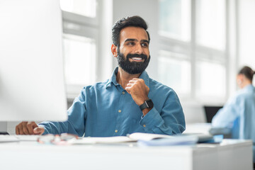 Aufkleber - Cheerful indian male office worker working on computer and smiling looking aside, sitting at workplace in office