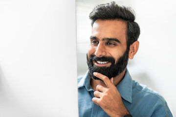 Aufkleber - Happy indian businessman working on computer in office, looking at monitor and smiling, touching chin, closeup shot