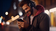 Smiling man holding mobile phone listening music at night outdoors, using smartphone doing chat in app, tech on cellphone, watching streaming tv videos online
