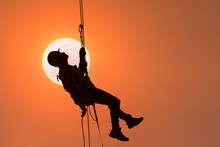 Silhouette Of A Construction Worker Hanging On A Safety Harness Against An Orange Sky At Sunset, Thailand