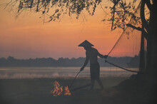 Silhouette Of A Fisherman Holding A Fishing Net And Making A Fire On Riverbank At Sunset, Thailand