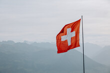 A Large Swiss Flag Evolves Against The Background Of The Swiss Alps, The Swiss Flag On Top Of The Mountain. The Wind Develops The Flag.
