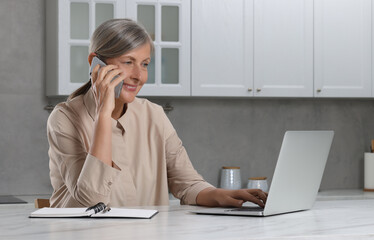 Wall Mural - Beautiful senior woman talking on phone while using laptop at white marble table in kitchen