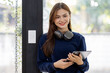 © David - Young adult happy smiling Hispanic Asian student wearing headphones standing indoor using digital tablet in university campus or at virtual office. College female student learning remotely.