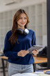 © David - Young adult happy smiling Hispanic Asian student wearing headphones standing indoor using digital tablet in university campus or at virtual office. College female student learning remotely.
