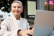 © Anatoliy Karlyuk - Portrait of happy confident senior lady freelancer in gray coat sitting outside at terrace with portable wireless laptop, working online, happy and proud with herself after finishing work