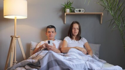 Family in bed unsatisfied wife taking remote control and choosing tv program while her husband using smart phone ignoring her scrolling online browsing web pages.