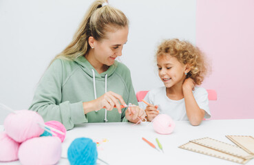 Mom teaches her daughter to knit. Mom and a little blonde girl are sitting at the table and crocheting.