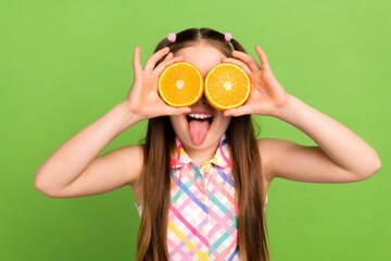 Portrait of little schoolgirl showing tongue out hold two halves orange fruit cover eyes isolated on green color background