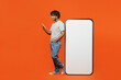 © ViDi Studio - Full body side profile view young Indian man he wears t-shirt casual clothes big huge blank screen mobile cell phone with workspace mockup area use smartphone isolated on orange red color background.