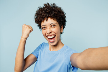 Wall Mural - Close up young happy woman of African American ethnicity wear t-shirt casual clothes doing selfie shot pov on mobile cell phone do winner gesture isolated on plain pastel light blue cyan background.