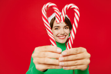 Wall Mural - Young fun woman wear green turtleneck Santa hat posing hold in hand look through candy canes in heart shape isolated on plain red background. Happy New Year 2024 celebration Christmas holiday concept.