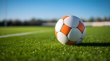 Close Up Of A Soccer Ball With White And Orange Patterns. Blurred Football Pitch Background