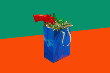 Blue Carton Bag On Red Surface With Tinsel, Red Toy Gun And Green Star Against Green Background