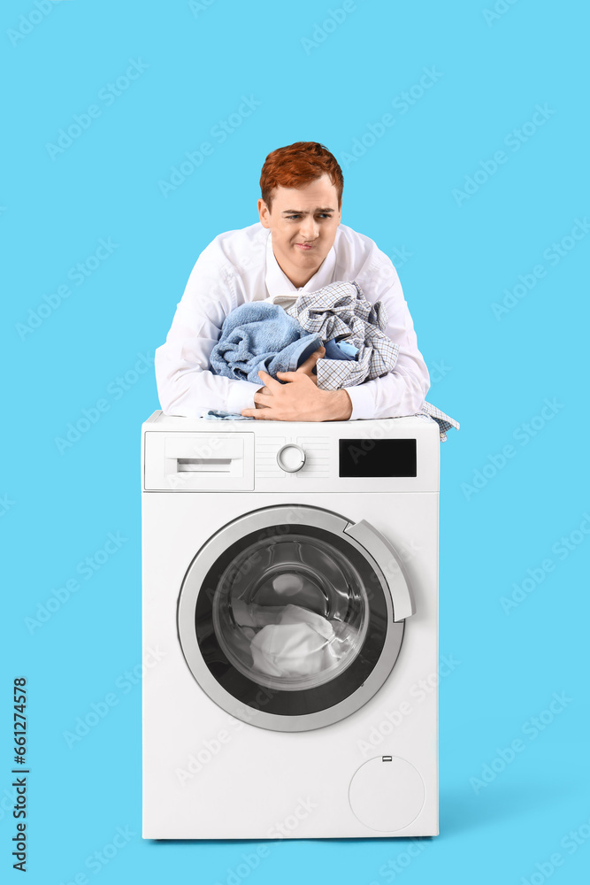 Displeased young man with laundry near washing machine on blue background