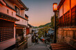 Kyoto City historic old town at sunrise with the warm blue sky in Japan, an empty downhill street with rustic wooden houses, cherry trees, and illuminated street light lamp