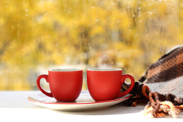 Wall Mural - a pair of red coffee cups on a saucer next to a blanket against the background of a window. autumn coffee break