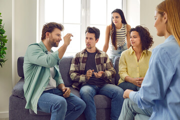 Wall Mural - Group of friends gather at home and spend time together. Several people sitting on the couch and talking. Bunch of students playing charades. Young man tells a story or explains a word to his pals