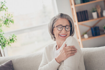 Sticker - Photo of positive adorable lady pensioner sitting on comfy sofa hugging herself enjoying autumn cozy day home indoors