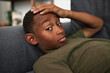 Indoor closeup image of bored lazy black kid boy lying on sofa with puzzled facial expression, thinking how spend leisure time without gadgets and internet, looking at camera touching head with hand