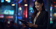 © Surachetsh - An Asian businesswoman, dressed in a sharp suit, utilizes a tablet against the backdrop of a cityscape aglow with the soft lights of the night.