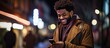 An African American man is using his phone on a city street at night while looking away
