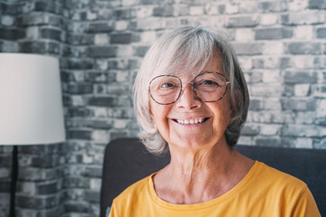 Smiling middle aged mature grey haired woman looking at camera, happy old lady in glasses posing at home indoor, positive single senior retired female sitting on sofa in living room headshot portrait.