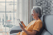 Smiling middle-aged Caucasian woman sit next to couch in living room browsing wireless Internet on tablet, happy modern senior female relax on ground at home using pad device, elderly technology