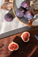 Wall Mural - Whole and cut fig fruits on plate and wooden cut board, on linen tablecloth with natural sunlight shadows. Healthy sweet autumn food, vitamin dessert for fall diet