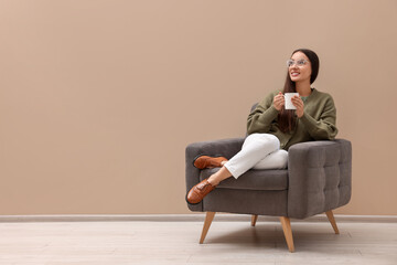 Canvas Print - Beautiful woman with cup of drink sitting in armchair near beige wall indoors, space for text