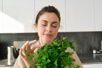 Portrait of attractive girl with bunch of parsley, eating fresh herbs and vegetables, cooking in the kitchen